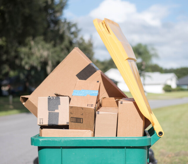 A green trash can full of boxes on a street.