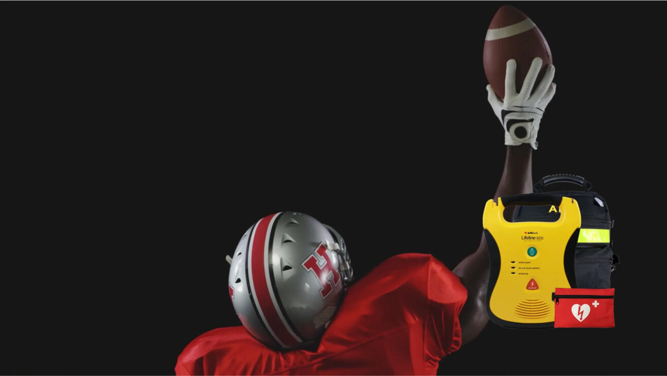 A football player in a red uniform, numbered 14884, reaches up to catch a football. A yellow automated external defibrillator (AED) and red first aid kit are visible in the foreground.