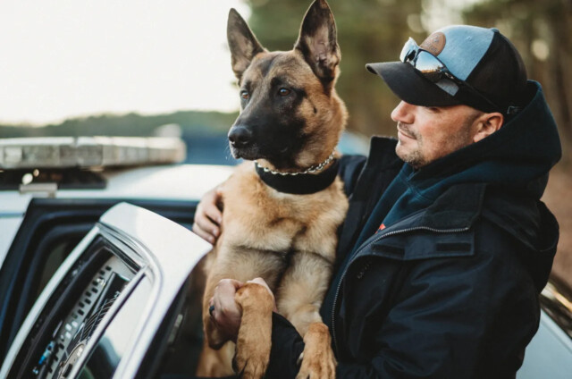 A man wearing a cap and sunglasses holds a German Shepherd dog next to an open police car door, with the number 14884 visible on the vehicle.