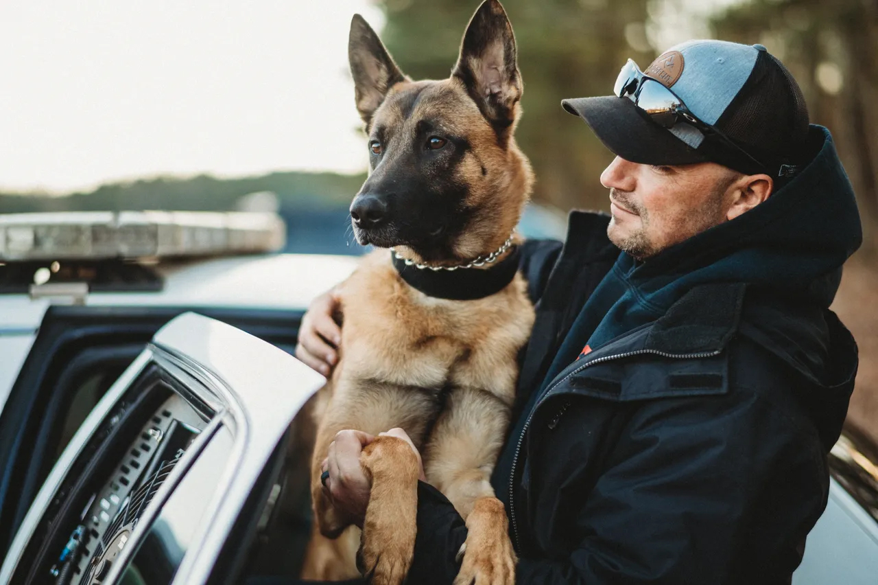 A man wearing a cap and sunglasses holds a German Shepherd dog next to an open police car door, with the number 14884 visible on the vehicle.