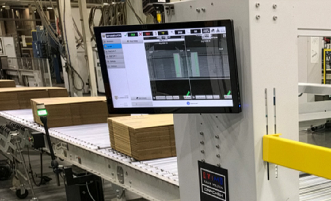 A computer monitor displays production data next to stacked cardboard sheets on a conveyor belt in a factory setting.