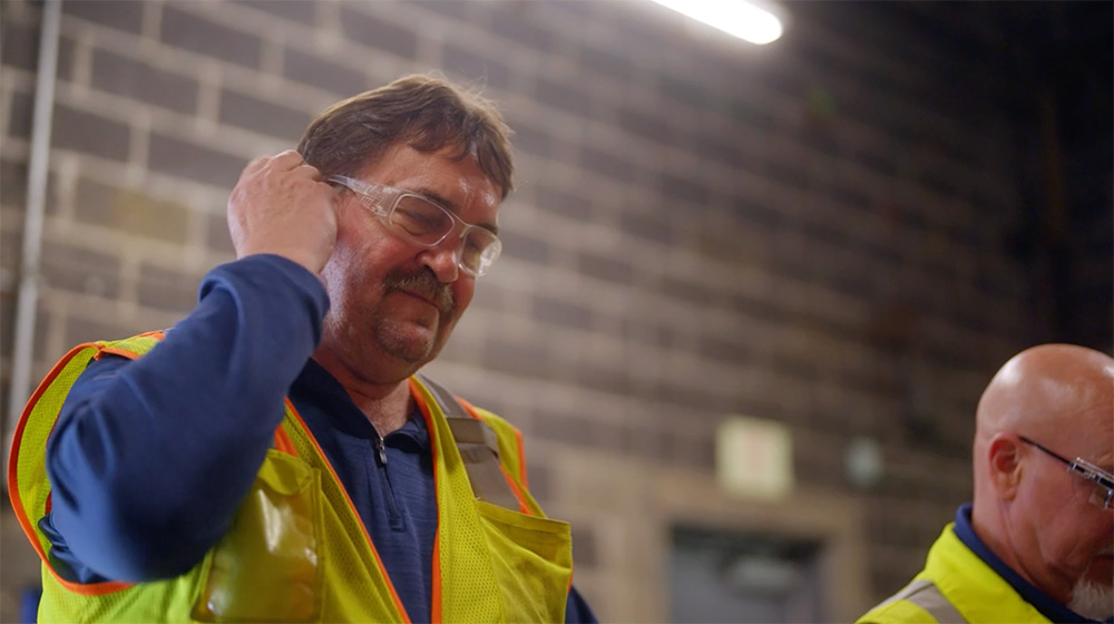 Two men in safety vests and glasses stand indoors; one adjusts his safety glasses. The background shows a brick wall and overhead lighting.